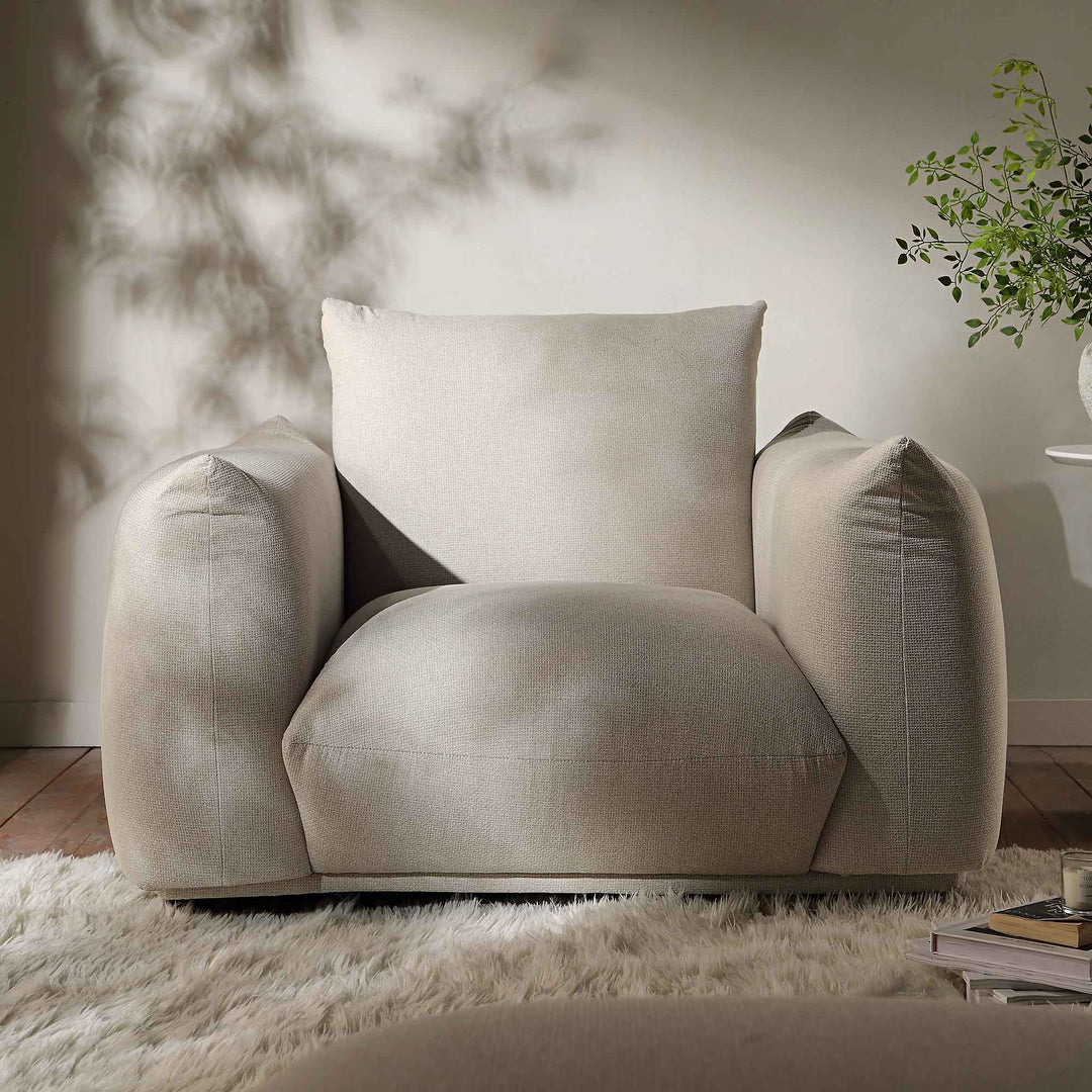 Beige armchair with a pillow in a room with a plant and books on a shelf.