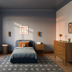 Load image into Gallery viewer, A styled children's bedroom featuring a bed with slate blue bedding and an orange accent pillow, set against a blue-gray vertically paneled wall. The room includes a wooden dresser, a patterned rug, and warm wall sconces.