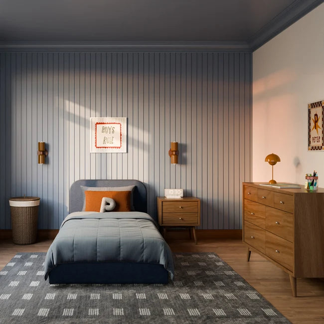 A styled children's bedroom featuring a bed with slate blue bedding and an orange accent pillow, set against a blue-gray vertically paneled wall. The room includes a wooden dresser, a patterned rug, and warm wall sconces.