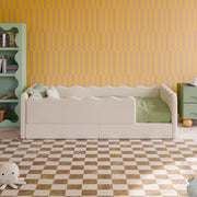 A children's bed with a cream upholstered frame and green bedding, styled in a room with a yellow wavy wall, a mint green bookshelf, and a checkered floor.