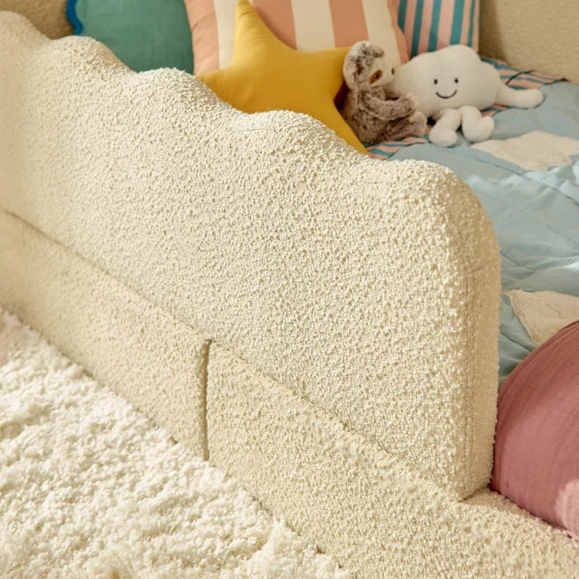 A close-up of a textured cream boucle or sherpa bed headboard in a child's bedroom setting, styled with colorful pillows and plush toys.