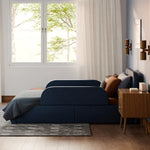 Load image into Gallery viewer, A navy blue upholstered bed with built-in head and foot guards, styled in a modern bedroom with a wooden nightstand, textured wall, and a window looking out to trees.