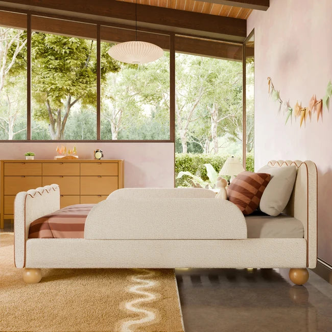 A mid-century modern upholstered bed with a cream bouclé headboard and footboard, styled in a bedroom with a pink wall, wood dresser, and large windows showing a green outdoor view.