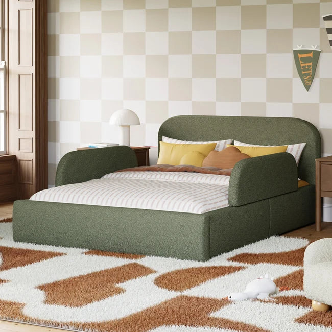 An upholstered bed frame in sage green fabric, styled in a child's bedroom with a checkered wall, patterned rug, and yellow accent pillows.