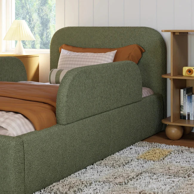 Green upholstered bed with brown and white pillows in a room with a window and bookshelf.