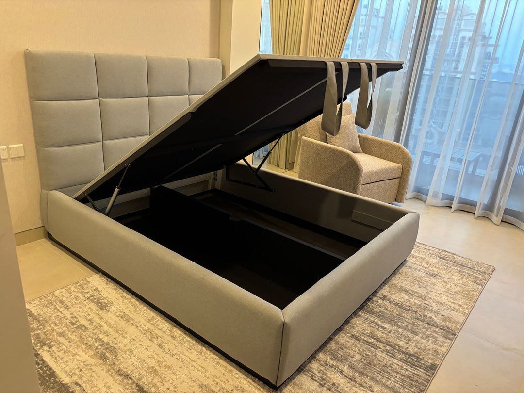 Gray upholstered bed with storage compartment in a room with curtains and a chair.